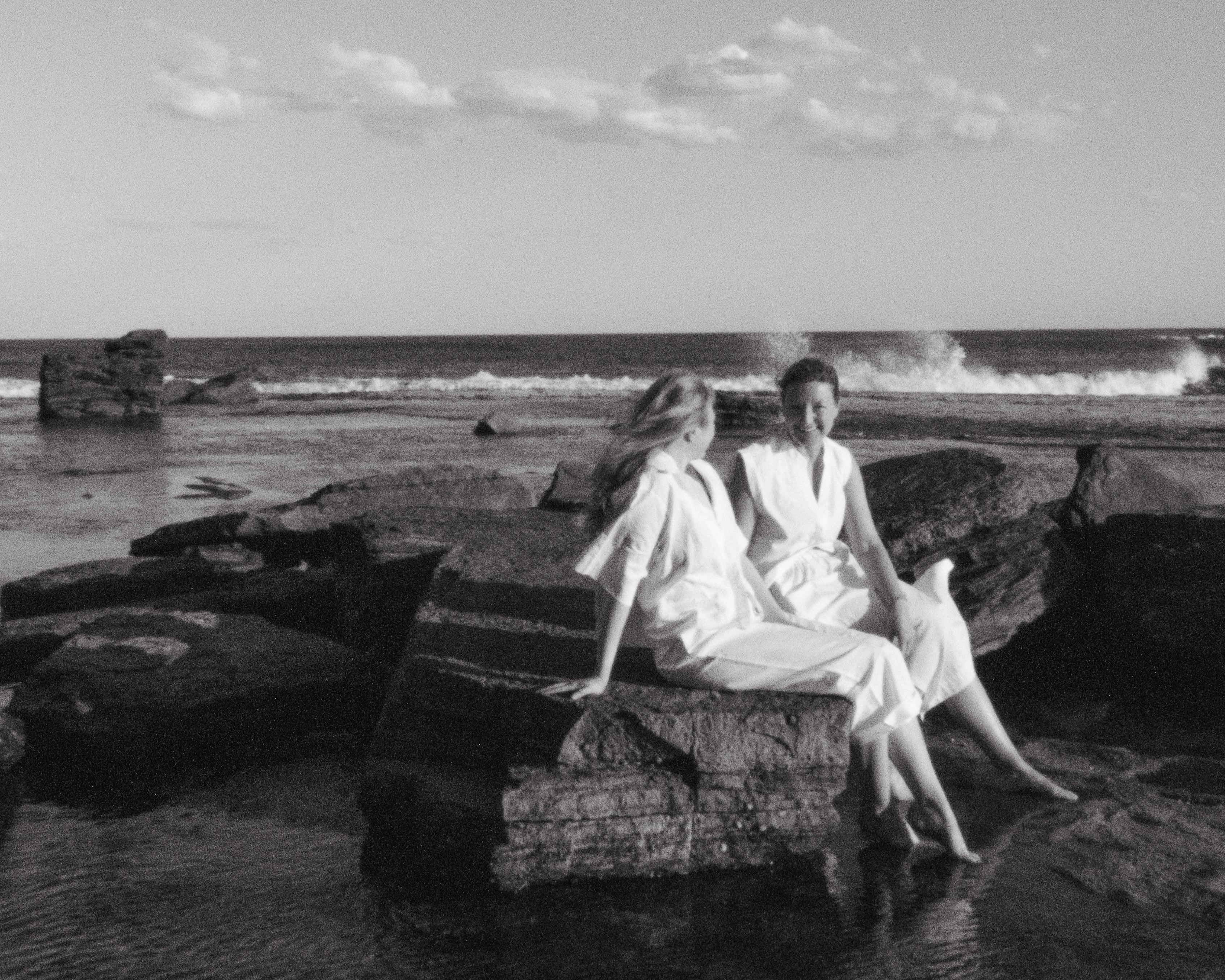 Two people sitting on rocks by the ocean with a minimal background
